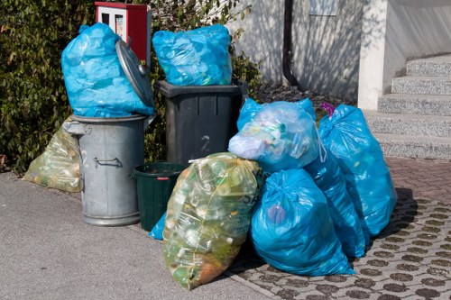 A collection of several overflowing black and grey wheelie bins and plastic rubbish bags placed on a paved area outside a building, with some bags and bins stacked or leaning against each other. The bags are mainly blue and transparent, revealing mixed waste inside, including food waste, plastic bottles, and paper. The scene is set outdoors, possibly on a driveway or pavement near a residential property, with greenery visible in the background and a section of concrete staircase on the right. The image depicts waste awaiting collection, illustrating local rubbish disposal and clearance services offered by House Clearance Slough, serving areas around Slough postcode and nearby towns.
