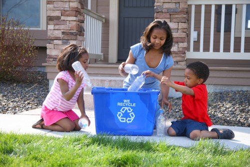 A young woman with medium skin tone and shoulder-length wavy hair, wearing a light blue sleeveless top, stands outdoors in front of a brick house with a dark grey front door and white railings, placing empty plastic bottles into a large blue recycling bin marked with the recycling symbol and the words 'We Recycle'. Sitting on a grey mat on a lush green lawn, two children with darker skin tones and dark hair are engaged in the activity; a girl with a pink and white striped top and pink skirt holding a plastic bottle close to her mouth, and a boy in a red t-shirt and dark shorts, reaching toward the bin with a plastic bottle in hand. The scene suggests an environmentally conscious activity, possibly related to rubbish or recycling collection, in a residential area near Slough. The background includes a garden bed with small shrubbery and a gravel space beside the path, emphasizing the outdoor setting. The overall atmosphere appears bright and sunny, with natural daylight illuminating the scene, supporting the context of rubbish management and recycling efforts led by House Clearance Slough.
