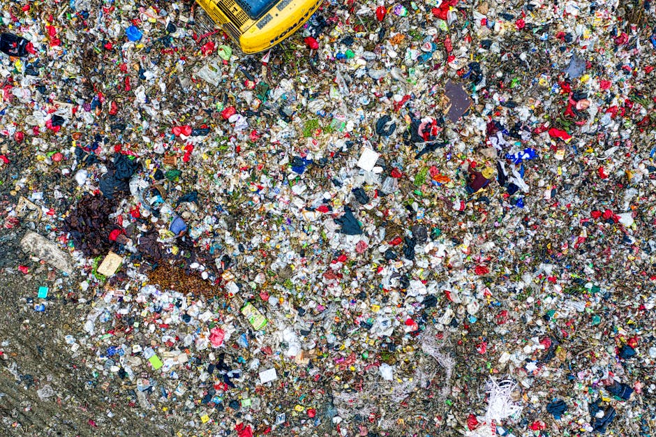 A collection of overflowing rubbish bins and assorted waste items is situated on a paved area adjacent to a roadside. The waste includes large black and black with black marked lids, a red bin, and a 
